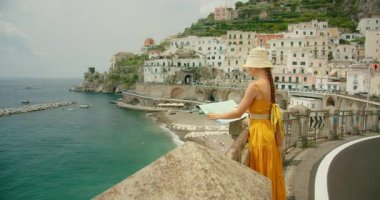 Bir İtalyan kasabası olan Atrani 'ye bakan bir sahil yolunun haritasını inceleyen bir kadın. Turist, Amalfi Sahili, İtalya, güneşli bir günde mimariye manzara açısından hayranlık duyuyor.
