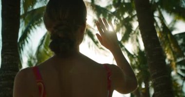 Kadın, tropikal plajda palmiye ağaçlarının altında dururken güneşi engellemek için elini kaldırır..