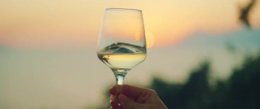 Yaz kıyı şeridinde gün batımını yansıtan bir şarap kadehi tutan kişi.