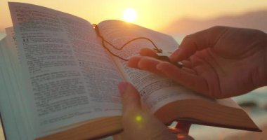 Gün batımında, ışık sayfaların üzerinde parlarken, kumsalda açık bir İncil ve haç kolye tutuyordu..