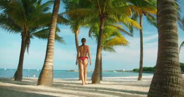 Kadın, tropik sahilde, açık mavi gökyüzünün altında, palmiye ağaçları arasında yalınayak yürüyor..