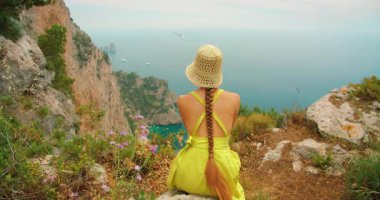 Yazlık elbiseli kadın uçurumun kenarında oturmuş Capri 'de güneşli bir günde denizi seyrediyor..