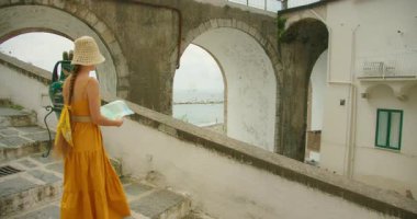 Sarı elbiseli kadın elinde kağıt haritayla Atrani 'nin yaz sokaklarını keşfediyor..