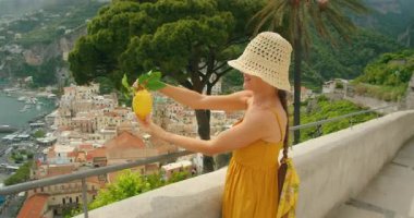 Gülümseyen kadın elinde büyük bir limon olan Sfusato Amalfitano ile arka planda Amalfi kasaba manzarası olan.