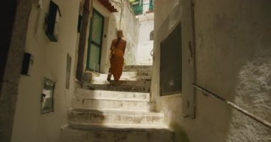 Yazlık elbiseli ve şapkalı bir kadın Amalfi caddesinde beyaz taş merdivenlerden yukarı çıkıyor..
