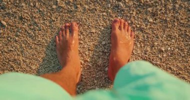 Çıplak ayaklı adam altın günbatımında kumsalda ıslak çakıl taşlarının üzerinde duruyor..