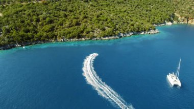 Bir jet ski derin mavi denizde kavis çizerken, yat ormanlık bir sahilde dinleniyor..