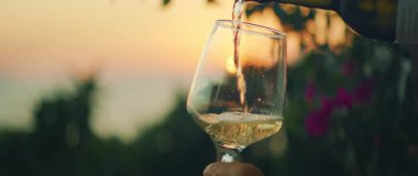 Gün batımında, arka planda deniz ve bahçe olan beyaz şarap kadehlerine dökülüyor..