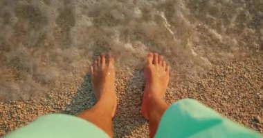 Sığ deniz suyunda ayaklarının altında çakıl taşlarıyla duran bir adam..