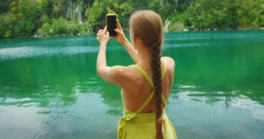 Hırvatistan 'ın Plitvice Lakes Ulusal Parkı' nda yeşil göl kenarında akıllı telefonla fotoğraf çeken kadın.