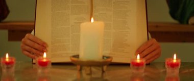 Eller, yansıtıcı sunaktaki yedi mumun arkasında açık bir İncil 'i tutar..