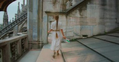 Bir kadın yürüyor ve izleyicileri mermer çatı terasında akşam karanlığında gotik mimariyle onu takip etmeye davet ediyor..