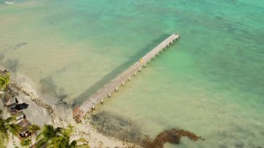 Güneşli bir günde, turkuaz tropikal denizin üzerinde uzun tahta bir iskelede yürüyen kişi..