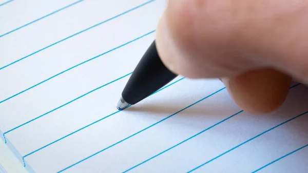 close up of right hand writing with black pen on blank white paper with blue lines as writing, note, office, message, information, work concept with copy space and selective focus.