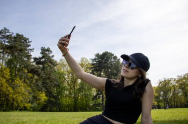 Siyah bluzlu ve siyah şapkalı bir kadın çim tarlasında oturur ve akıllı telefonuyla fotoğraf çeker..