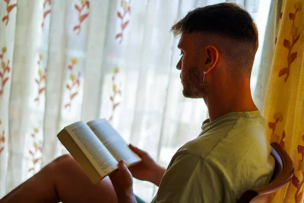 Man with half dye blonde hair reading a book close to the window