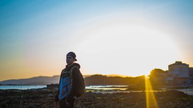 Gün batımında plajda ciddi poz veren genç adam. Şapka ve sırt çantası takıyor.