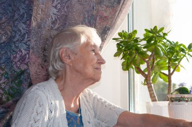 Evde pencerenin yanında oturan yaşlı yalnız kadın.