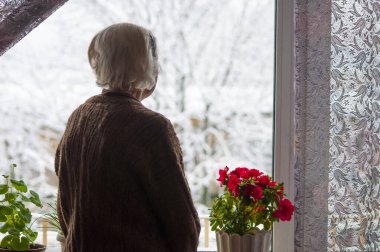 Yaşlı yalnız kadın evinin penceresinin yanında duruyor ve kışın bakıp hayal kuruyor.