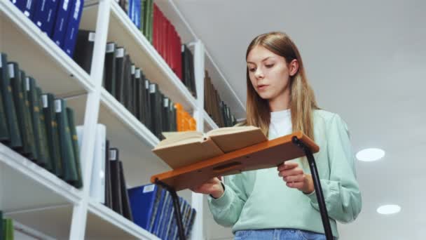 図書館ではしごに立って本を読んでいる白人の十代の少女 ページをめくる 面白いプロットを楽しんで 低角度の学生は 小説を楽しんで 新しい科目を学ぶ 知識の概念 — ストック動画