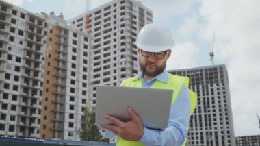 Dikkatli sakallı bina müfettişi, sıkı şapka ve güvenlik yeleği giyiyor dizüstü bilgisayara not alıyor, arka planda çok katlı evler var. Düşük açılı yay adam inşaat alanını inceliyor. Kontrol kavramı