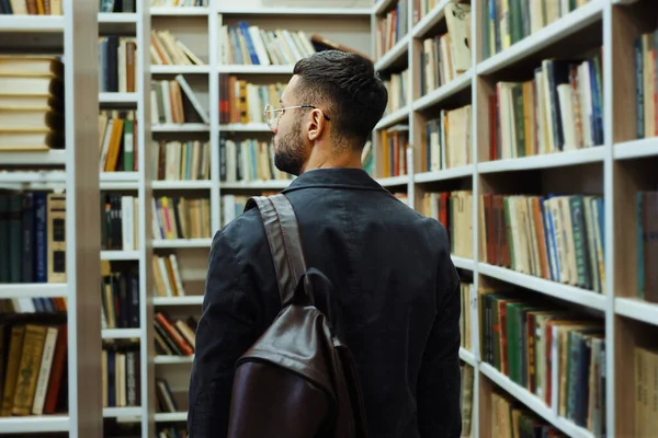 stock image Man wearing jacket with backpack on shoulder walking past bookcases. Following shot college student searching for necessary text books or literature in library. Concept of education