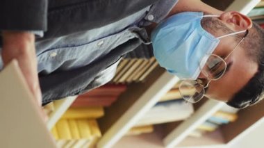 Gözlüklü ve koruyucu maskeli bir adam kitapçıda oturuyor ve kitap okuyor, arka planda kitaplıklar, düşük açı. Kütüphanede okuyucu. Eğitim kavramı