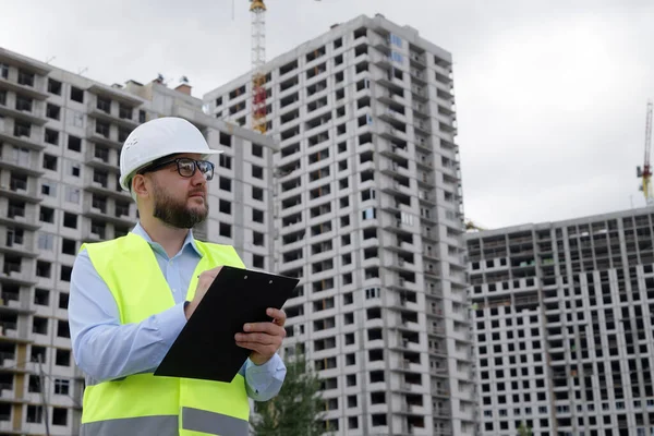 Bearded building inspector wearing hard hat and safety vest making notes on paper, multistory houses on background. Low angle man inspecting construction site. Concept of control