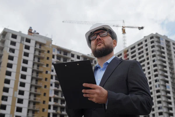 Bearded building inspector wearing hard hat and suit jacket making notes and talking to himself, multistory houses on background. Low angle man inspecting construction site. Concept of control