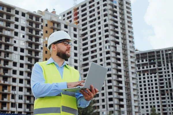 Bearded building inspector wearing hard hat and safety vest making notes on laptop, multistory houses on background. Low angle man inspecting construction site. Concept of control