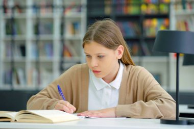 Smart girl in casual wear sitting at desk and writing in exercise book, bookshelves on blurred background. Pupil doing assignment in class. Concept of education