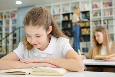 School girl sitting at desk and laughing while reading funny book, another pupil behind her, teacher standing by bookshelves and choosing book on blurred background. Concept of education