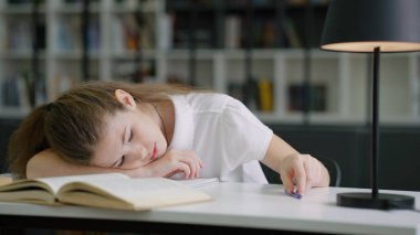 Masada uzanan ve kendini eğlendirmek için kalemle oynayan liseli kız, okulda sıkıcı dersler alan öğrenci, arka planda bulanık kitaplıklar. Eğitim kavramı