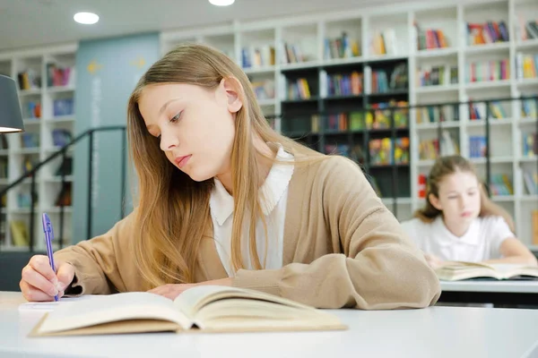 Uzun saçlı, günlük giysiler giyen, çalışma masasında oturan ve egzersiz kitabına yazan bir kız öğrenci daha bulanık arka planda. Çocuklar ödevlerini yapıyor. Eğitim kavramı