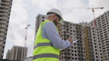 Miğfer ve güvenlik yeleği giyen sakallı adam dosya klasörü tutuyor ve inşaat halindeki çok katlı binalara karşı notlar tutuyor. Düşük açılı müfettiş olay yerinde yürüyor ve kalitesini kontrol ediyor.