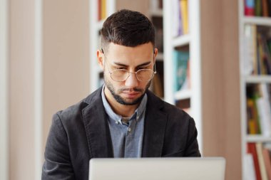 Gözlüklü genç adam kütüphanede dizüstü bilgisayarda çalışıyor, internette geziniyor, araştırma yapıyor, uzaktan çalışıyor, çevrimiçi çalışıyor, arka planda bulanık kitaplıklar var. Eğitim kavramı