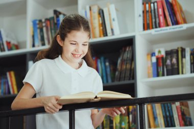 Metal parmaklıklara yaslanmış, kitap okuyan, arkasında kitaplıklar olan beyaz tişörtlü mutlu bir kız. Kütüphanede edebiyattan keyif alan okul çocuğunun portresi. Eğitim kavramı