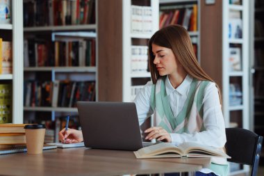 Kütüphanede dizüstü bilgisayarla çalışan, kağıt üzerinde notlar alan, masanın üzerinde duran kitapları açan, yanında karton bardakla uzun saçlı bir kız öğrenci. Eğitim kavramı