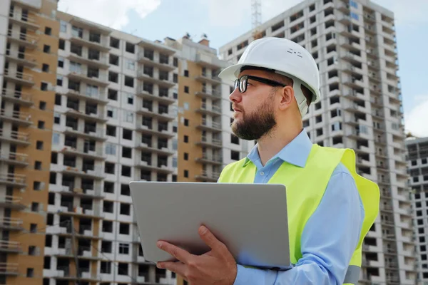 Watchful bearded building inspector wearing hard hat and safety vest making notes on laptop, multistory houses on background. Low angle man inspecting construction site. Concept of control