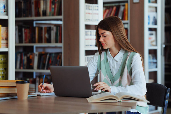 Студентка с длинными волосами, работающая в библиотеке на ноутбуке, делающая заметки на бумаге, открывающая книги, лежащие на столе, картонная чашка рядом. Концепция образования