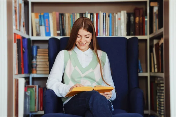 Young woman with long hair in casual clothes sitting in cozy armchair and reading book and laughing at funny episodes, bookshelves on blurred background. Concept of literature