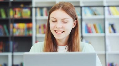 Kütüphanede dizüstü bilgisayarında video görüşmesi yapan gülümseyen genç kız, webcam ile konuşuyor, telekonferans ya da zoom etkinliği yapıyor, arka planda bulanık kitaplıklar olan kitaplıklar. Online rota. Uzaklık öğrenimi