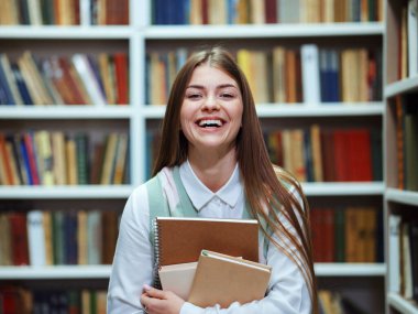 Kitaplıkların arasında duran, elinde kitap yığınıyla kameraya gülen uzun saçlı genç bir kadın. Kütüphanedeki mutlu üniversite öğrencisinin portresi. Eğitim kavramı