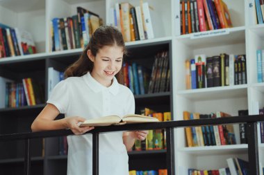 Cheerful girl wearing polo shirt standing by bookshelves and reading book, flipping pages. Portrait of child enjoying literature in book shop or school library. Concept of education