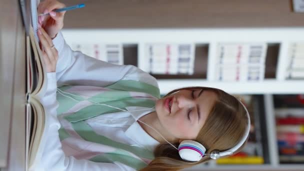 垂直屏风 女生坐在图书馆里 做作业 用耳机听音乐 站在她面前的笔记本电脑 在书中学习的年轻女子 教育的概念 — 图库视频影像