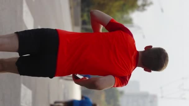 縦型スクリーン 赤いTシャツを着たスローモーションの男が スマートフォンを手に空の街の道をマラソンしている スポーツイベントでのシュートランナーに続く — ストック動画