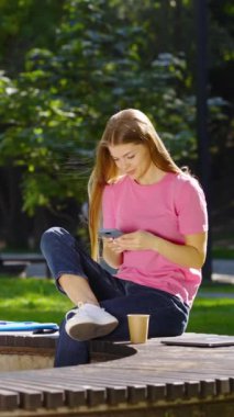 Dikey Ekran: Uzun saçlı, günlük kıyafetler giyen, yaz parkında bankta oturan ve akıllı telefondan sosyal medyada gezinen genç bir kadın. Aygıt kavramı