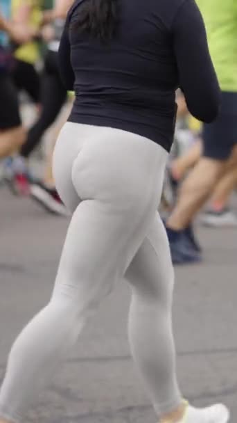 縦型スクリーン 黒いスポーツジャケットと白いレギンスを身に着けているスローモーション脂肪の女性は 彼女の手の中に他の人 スマートフォンと一緒に都市道路上で実行されます マラソン競技者の一部の見解 — ストック動画