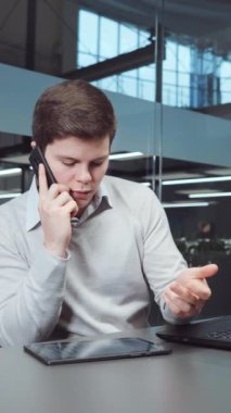 Süveter giyen genç adam ofiste çalışıyor, dizüstü bilgisayar kullanıyor, telefonla konuşuyor ve tabletten ekranları aşırıyor. İş yerindeki mali danışman