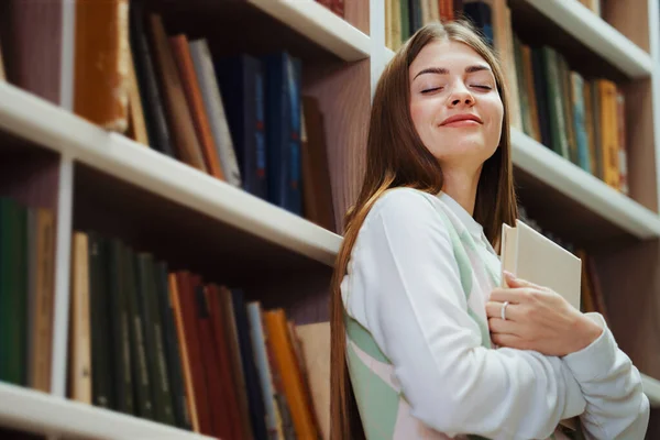 Young female with long hair leaning on bookcase and closing book with happy smile on face and starts dreaming with eyes closed. Low angle reader enjoying romantic novel in library